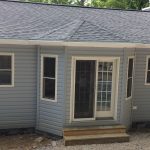 A small house with a gray roof and white trim is sitting on top of a gravel lot.